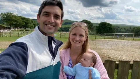 Caroline Lambert A man wearing a blue and white jumper smiles at the camera as he stands in a field. He is next to a woman wearing a pink cardigan as she holds a baby in a blue outfit.