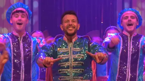 Three people in a pantomime on stage. They are singing and smiling. A man with short dark hair in the centre is wearing a green sparkly coat with gold buttons. The two people either side of him are wearing blue sparkly coats with purple stripes running vertically down the centre