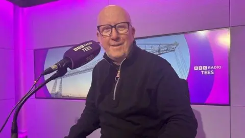 Andy McDonald is sitting in a studio with a large microphone in front of him with the words Radio BBC Tees on it and behind him the words appear on a screen showing the Transporter Bridge. He is a man in his 60s with not much hair, glasses and wearing a black top with quarter length zip.