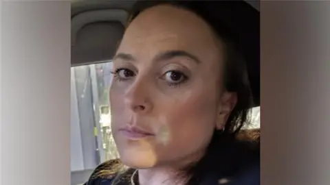 A woman with dark shoulder-length hair who appears to be sitting in a car looks at the camera. She has dark eyes and a small nose stud.