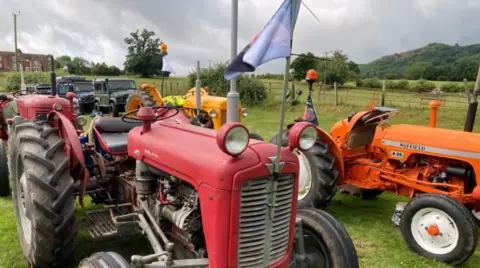A red tractor has a pole with a flag fixed to the front. The flag has a picture of Harry on the opposite side. There are other vehicles around the tractor in different colours.

