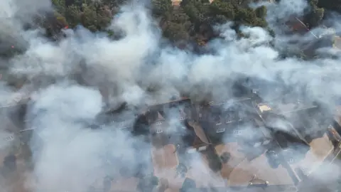 PA Media An aerial photo of grey smoke hanging above houses with trees behind.