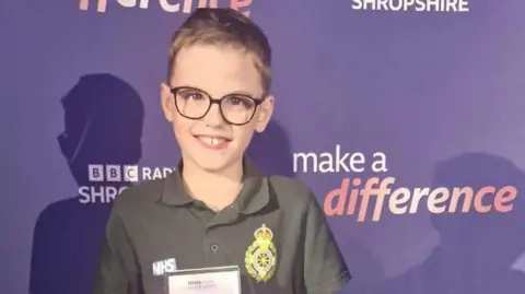 Family photo A boy with short brown hair, glasses and a green top with a yellow badge in front of a purple background