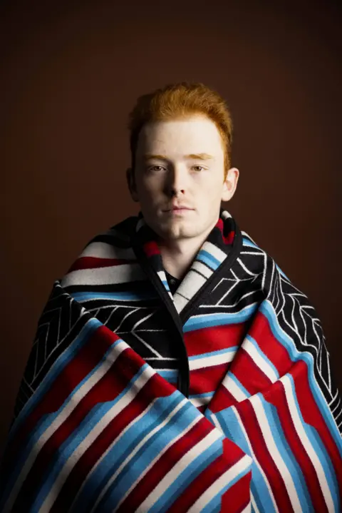 Kieran Dodds Studio portrait of a person with short red hair wrapped in a patterned blanket featuring red, blue, black, and white stripes and geometric designs. The person faces the camera against a dark brown background, with the blanket draped around the shoulders and covering most of the body.