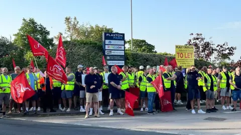 A large group of people gathered on a roadside outside an industrial estate taking part in a trade union picket, wearing high-visibility yellow vests. A number are holding red Unite the Union flags. Behind the group is a blue sign reading: PLAXTON PARK.
