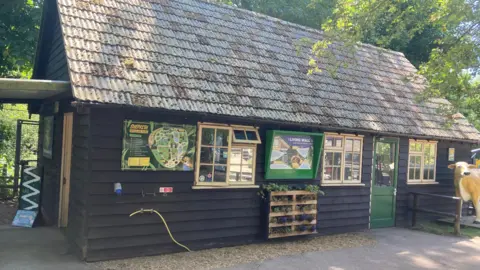 Josh Lovatt/BBC A single-storey wooden lodge is surrounded by trees and has several information boards, including a map of Shepreth Wildlife Park, on the outside walls. There is a green door to the right and there is also a statue of a cow.