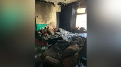 Paul Fosh Auctions The inside of the same terraced house in another room, with new debris including a burnt and dirt covered sofa and peeling burnt wallpaper 