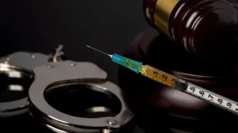 A stock image that shows handcuffs, a gavel and a syringe