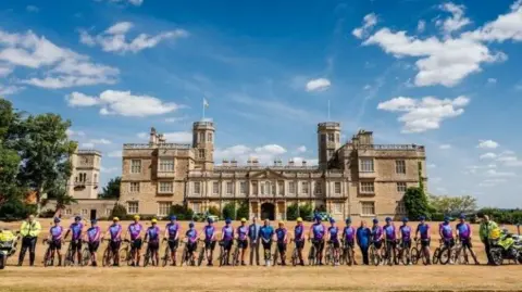 Northamptonshire Police A group of people wearing lycra cycling gear are standing in a row with bicycles. They are flanked at each end by a police motorcycle officer standing next to their motorcycle. The group picture has been taken in front of an elaborate brick building with towers and flags.