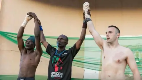 Kayode Oluwa A man stands in the centre of two other men holding their arms up in the air. The man in the middle is wearing a black t-shirt which says 'African warriors' on it. The other two men are topless.