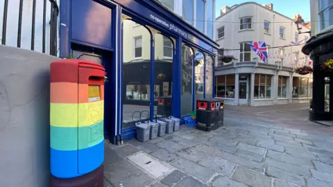 A Guernsey post box painted five different rainbow colours and a blue shop with large windows with food bins and a public bin next to it.