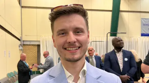 Nicola Haseler/BBC Liam Morrell-Phillips with short brown hair and beard, wearing a light purple jacket and yellow shirt, with people behind him at an election count. The room has yellow walls and beige curtains.