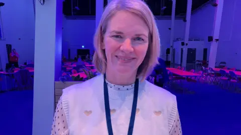 Tom Ingall/BBC A woman with blonde bobbed hair wearing a white blouse and a cream tank top with hearts on. She is smiling and wearing a blue lanyard. There are tables and chairs in the background. 