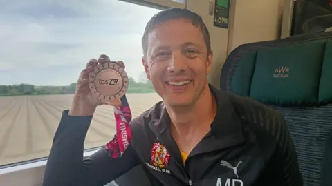 Michael Barrett Michael Barrett smiles at the camera holding his London Marathon Medal. He is sat on a train, there is a field behind him through the window. 
