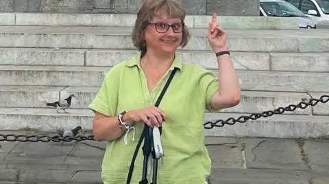 Jules wearing a green blouse, and glasses with walking sticks in her hand. She is smiling at the camera and pointing up.