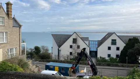 The view from the garden with Godrevy lighthouse in the distance across the sea. In the foreground beyond a boundary wall is a digger at work, a site office container and a white van. Across the road is a large building with an atrium between its two halves.