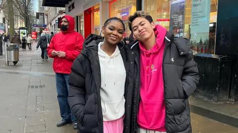 Thando Sibenke wears a black coat and white hooded top standing next to her friend on a street in Birmingham