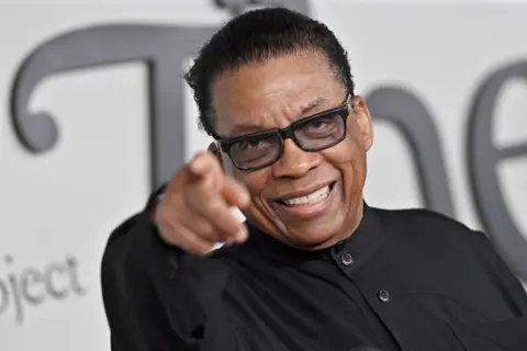 Getty Images Herbie Hancock apontando direto para a câmera. Ele está usando óculos pretos e uma camisa preta sem gola.