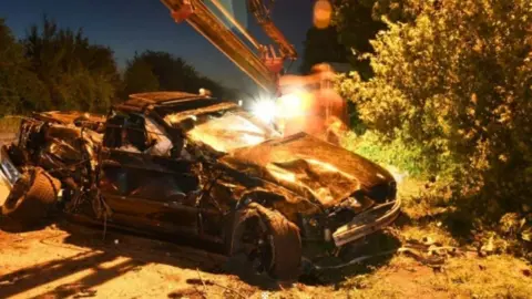 A photo of the wreck of black BMW car, taken at night. The car is a mangled wreck with the front bar of a crane visible overhead. The car's rear is nearly completely ripped away.