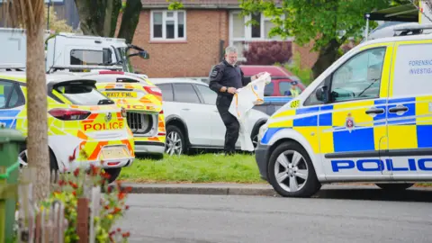 PA A policeman wearing all black is holding some white overalls. There are a number of police vehicles parked up on some grass.