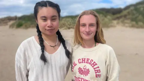 Revati Campbell and Maya Spanabel smile for the camera while standing on Irvine beach - Revati has dark hair with pigtails and a white/cream top on. Maya has blond hair and a yellow top on.