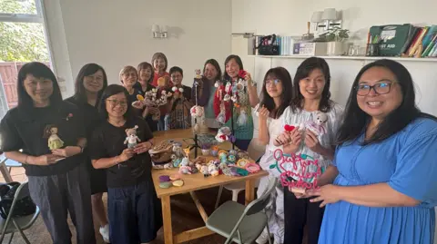 12 members of the group stand around a table showing off what they have created, from crocheted children's teddies, to coasters and poppies
