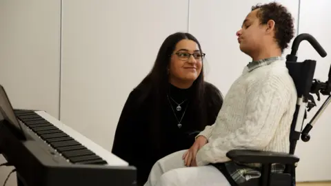 Karima Motala sits beside her son Jaden, who is in a wheelchair next to a keyboard, looking towards him in a bright room.