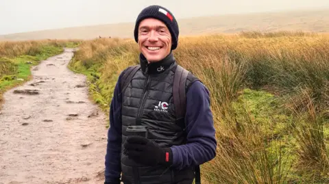 JC Explores Jay smiling looking at the camera. He is standing on a muddy walking path on a mountain. Jay wears a long sleeve navy top, a navy hat, a black gilet, black gloves and holds a takeaway coffee in his left hand.