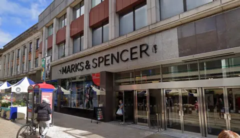 The grey concrete department store has a large Marks and Spence sign on the front with shoppers walking in and out