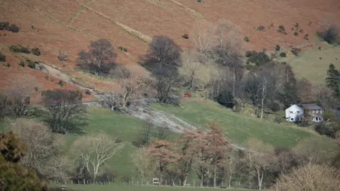 BGS A large hill slope with trees and brown and green vegetation. In the middle of the slope is a crevice with debris coming down the slope. A house sits to the right of it at the bottom of the hill.