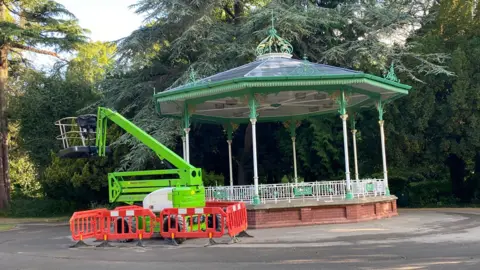Durham County Council The green and white bandstand structure on a base of red bricks, which sits in the middle of a park. There is a green cherry picker next to it and a thick grouping of trees stands in the background.