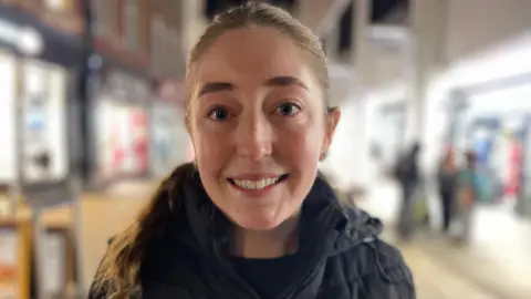 Laura Foster/BBC Emily Brown is smiling directly at the camera. She has light brown long hair tied back in a ponytail. She is wearing a black padded jacket and top. The background of shops is blurred.