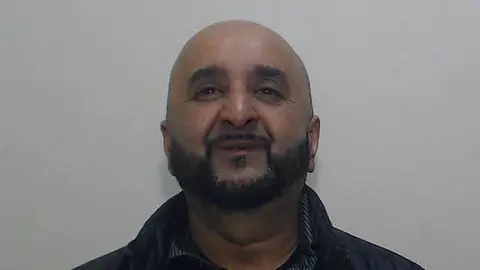 Police custody shot of Ajaz Ahmed who is bald with a black beard wearing a black jacket and black shirt