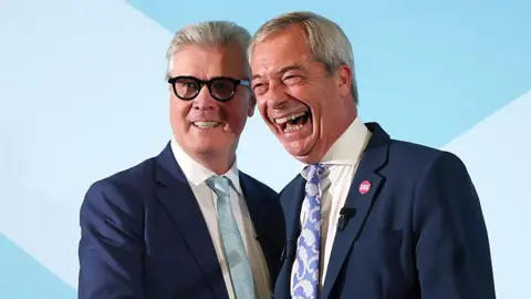 Getty Images Malcolm Offord and Nigel Farage standing close together on a stage. They both have short, grey hair and are wearing blue suits and cream or white shirts. Offord has dark-rimmed glasses and is wearing a light blue tie. He is smiling while Farage, who is laughing, is wearing a paisley-patterned blue and white tie. 