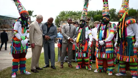 PA Media King Charles III wears a beige suit and speaks to a man in a grey suit. Standing in a row beside them are Gombey dancers in neon outfits with tassels, masks and tall headdresses