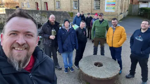 For Men To Talk A group of men, in outside coats, at Jordans Mill, standing by a large mill stone. They are all looking at the camera and smiling.