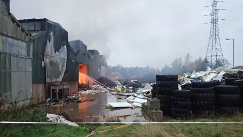 Provided by the Environment Agency Wide view of the NWR site with the fire burning inside one unit, with water being poured onto the fire, tyres piled up to one side and what looks like an image of a giant chicken painted on the wall.