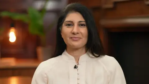 Aliya is wearing a cream, buttoned blouse and has long, black hair. She is smiling at the camera and is sat in a room with oak-lined walls. In the background, blurred, are a desk, lamp and plant. It is a head and shoulders shot of her.