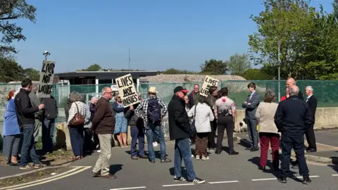 Campaigners gather in front of the site of the proposed warehouse on a sunny day. Three of them hold signs that say "lives not lorries."