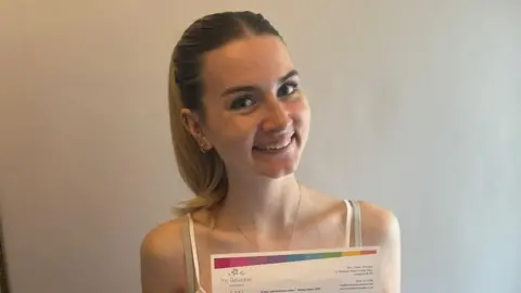 Lily-Rose Williamson Lily-Rose Williamson, a young woman with long blonde hair tied back in a ponytail, smiles for the camera in front of a plain white wall. She is holding a sheet of paper with her A-level results printed on it, the top of which is visible in the image.