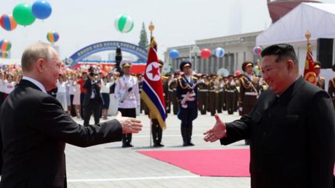 Mr Putin and Mr Kim smile and reach out towards each other to shake hands, with a parade in the background