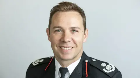 A picture of Mark Arkwell, who has short, brown hair, wearing his Royal Berkshire Fire and Rescue Service uniform, which includes a black jacket, black clip-on tie and white shirt.