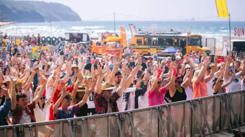 Hundreds of people stand at the barrier of a music festival on a beach with their hands in the air. It is a sunny day and you can see the sea in the background.