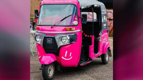 A pink tuk-tuk is parked on concrete outside. It is covered in branding and these words are printed: Rosie the little pink tuk tuk