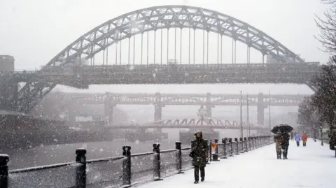 PA Media Snow falls on the Quayside in Newcastle. People in winter coats and hats make their way down the snowy path next to the Tyne river, with the arched Tyne Bridge rising in the sidtance.