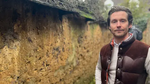 Sam Falco looking into the camera, standing next to a mud wall. He has brown hair and is wearing a brown gillet and a beige jumper.