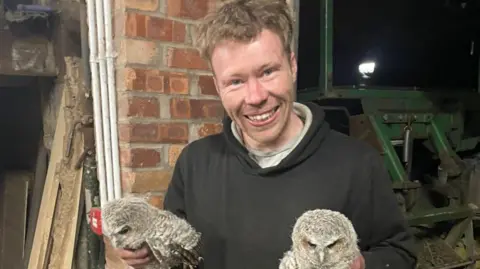 CPS Carruthers, who has short fair hair and is wearing a black hoody, holds two young grey owls. He is smiling with agricultural equipment behind him
