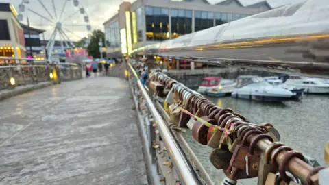 Image of padlocks along the handrail of Pero's Bridge. Most look rusty. Boats can be seen moored in the background. In the distance, the light of cafes and restaurants can been seen, with people walking along. A ferris wheel can also be seen.