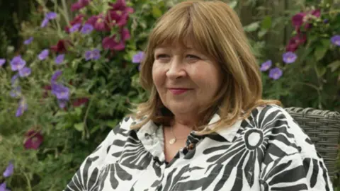 A woman in a black and white floral blouse sits on a garden chair. Flowers can be seen in the background.