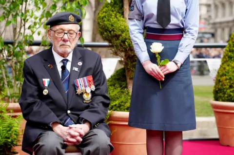 PA Media Second World War veteran Alan Kennett closes his eyes while sitting down wearing a suit with military badges on his lapel and a cadet hat, next to a person wearing a skirt holding a white rose, as they gather to watch the procession.